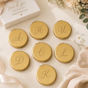 May include: Seven round gold-toned metal compact mirrors, each engraved with a unique cursive letter including A, H, U, D, C, L, and K. A white gift box is visible with the text Thank you for being my bridesmaid and a small heart icon.