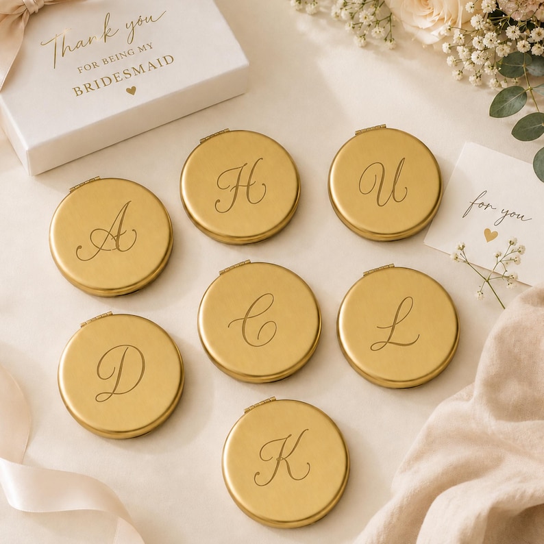 May include: Seven round gold-toned metal compact mirrors, each engraved with a unique cursive letter including A, H, U, D, C, L, and K. A white gift box is visible with the text Thank you for being my bridesmaid and a small heart icon.