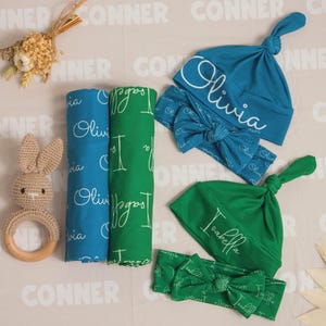 May include: A selection of baby accessories, featuring two swaddling blankets, two knotted hats, and two headbands. The blankets are blue and green, with the names "Olivia" and "Isabella" printed on them. The hats and headbands match the blankets.