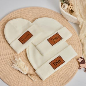 Op de afbeelding: Crèmekleurige gebreide mutsen met bruine leren naamlabels. De labels zijn voorzien van de namen "Hannah", "Grace" en "Erin". De mutsen zijn omgeslagen aan de rand. De achtergrond omvat een geweven mat en decoratieve elementen.
