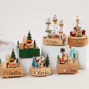 May include: A collection of wooden music boxes with various themes, including trains, Ferris wheels, and carousels. Each box features intricate details, miniature figures, and personalized names like "Emilia," "Lilly," and "Aria." The boxes are set on a white surface.