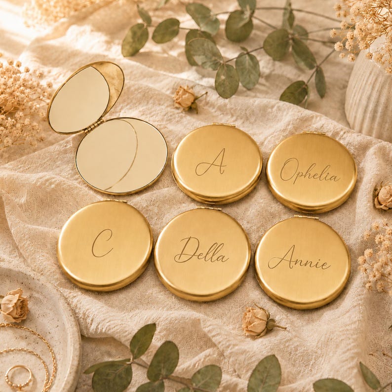 May include: A collection of round gold-toned metal compact mirrors with hinged lids. Five mirrors are closed, displaying engraved names or initials including A, C, Della, Ophelia, and Annie in a script font. One mirror is open, revealing two reflective surfaces.