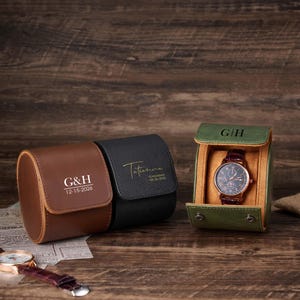 Può includere: Tre custodie per orologi in pelle marrone, nera e verde. La custodia marrone ha le iniziali "G&H" e la data "12-15-2026". La custodia nera ha il nome "Tatiana" e la data "06-26-2026". La custodia verde contiene un orologio.