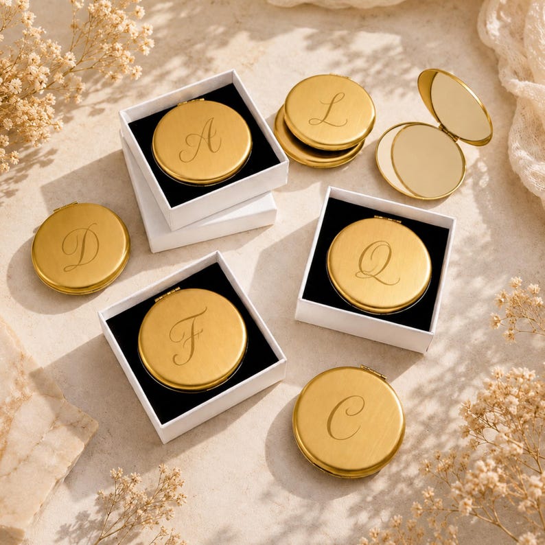 May include: A collection of round gold-toned metal compact mirrors, each engraved with a single cursive letter. Some mirrors are placed inside square white gift boxes with black inserts, while others are shown open to reveal the reflective surface.