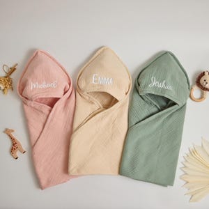Op de afbeelding: Drie babyhanddoeken met capuchon in roze, beige en groen. Elke handdoek is geborduurd met een naam: "Michael", "Emma" en "Joshua". De handdoeken zijn gemaakt van een zachte, getextureerde stof. Een houten giraf en een bijtring in de vorm van een leeuw liggen in de buurt.