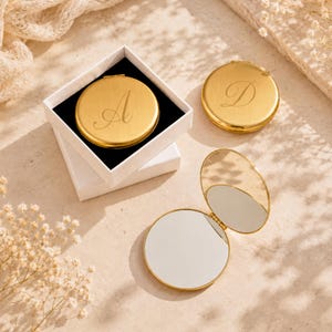 May include: Three round gold-toned metal compact mirrors. One mirror is inside a white square box and features the letter A engraved on the lid. A second closed mirror features the letter D. A third mirror is open, revealing two reflective surfaces.