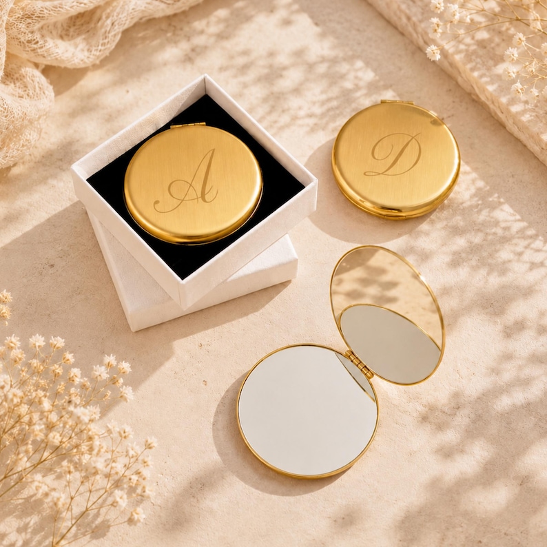 May include: Three round gold-toned metal compact mirrors. One mirror is inside a white square box and features the letter A engraved on the lid. A second closed mirror features the letter D. A third mirror is open, revealing two reflective surfaces.