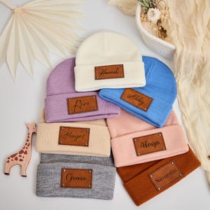Puede incluir: Varios gorros de bebé personalizados en colores crema, lavanda, azul, rosa, beige, gris y marrón. Cada gorro tiene un parche de cuero sintético marrón con un nombre en escritura cursiva. Se ve un juguete de madera con forma de jirafa.