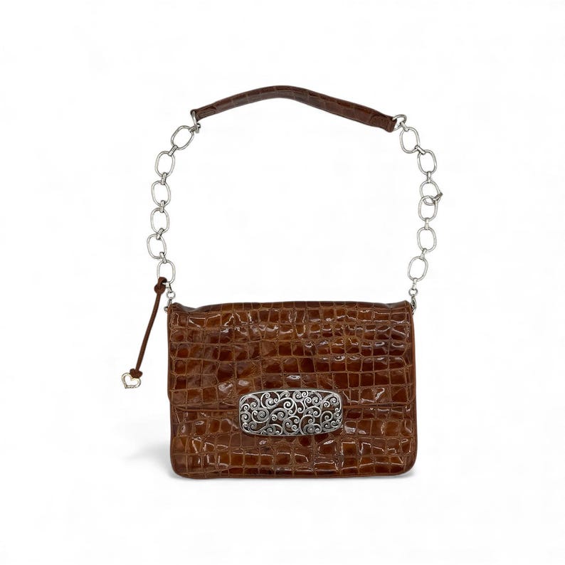 Puede incluir: Un bolso rectangular marr&oacute;n con textura de piel de cocodrilo. El bolso presenta un cierre decorativo plateado y una correa de cadena plateada con un asa de cuero marr&oacute;n. Cuelga un cord&oacute;n de cuero marr&oacute;n con un dije en forma de coraz&oacute;n.