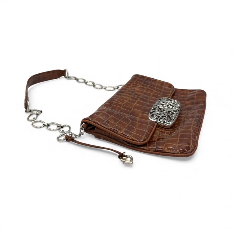 Puede incluir: Un clutch de cuero marr&oacute;n con una cadena plateada. El clutch tiene una textura de cocodrilo y un cierre plateado con un dise&ntilde;o ornamentado. Una peque&ntilde;a correa de cuero con un dije en forma de coraz&oacute;n cuelga de la cadena.