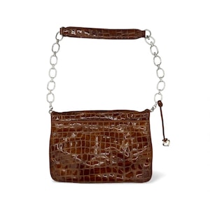 Puede incluir: Un bolso marr&oacute;n con textura de cocodrilo, una correa de cadena plateada y un peque&ntilde;o dije de coraz&oacute;n. El bolso tiene forma rectangular y un acabado brillante. El asa tambi&eacute;n es marr&oacute;n y texturizada.