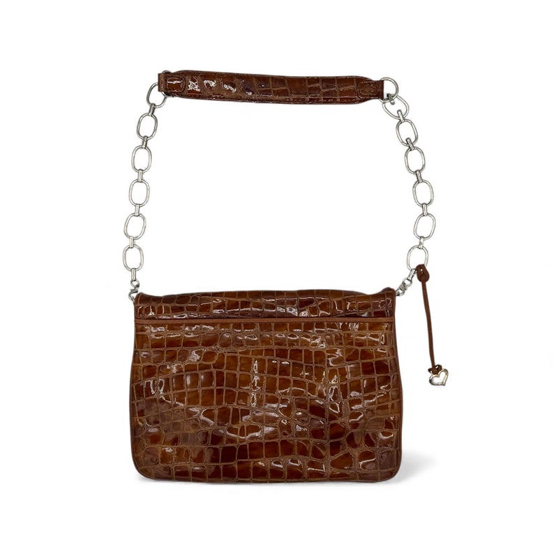 Puede incluir: Un bolso marr&oacute;n con textura de cocodrilo, una correa de cadena plateada y un peque&ntilde;o dije de coraz&oacute;n. El bolso tiene forma rectangular y un acabado brillante. El asa tambi&eacute;n es marr&oacute;n y texturizada.