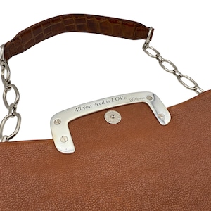 Puede incluir: Bolso de mano de cuero marr&oacute;n con un asa plateada que dice "All you need is LOVE" y un asa marr&oacute;n con textura de cocodrilo. El bolso tiene una correa de cadena plateada.