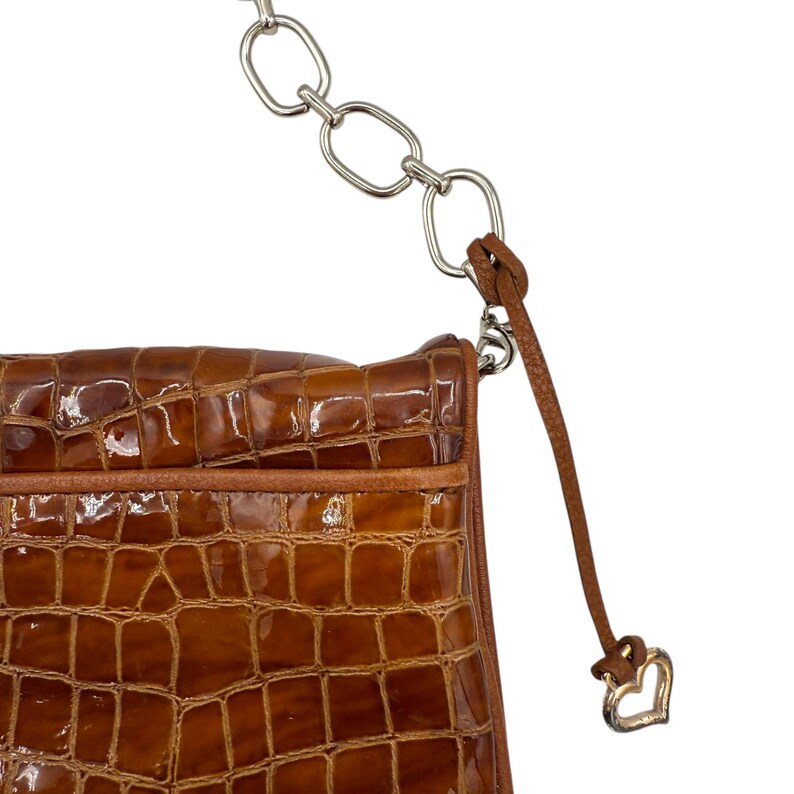 Puede incluir: Un bolso marr&oacute;n texturizado con una correa de cadena plateada. El bolso tiene un patr&oacute;n similar al de un cocodrilo y una correa de cuero marr&oacute;n con un dije de coraz&oacute;n plateado. El bolso es un bolso de hombro.