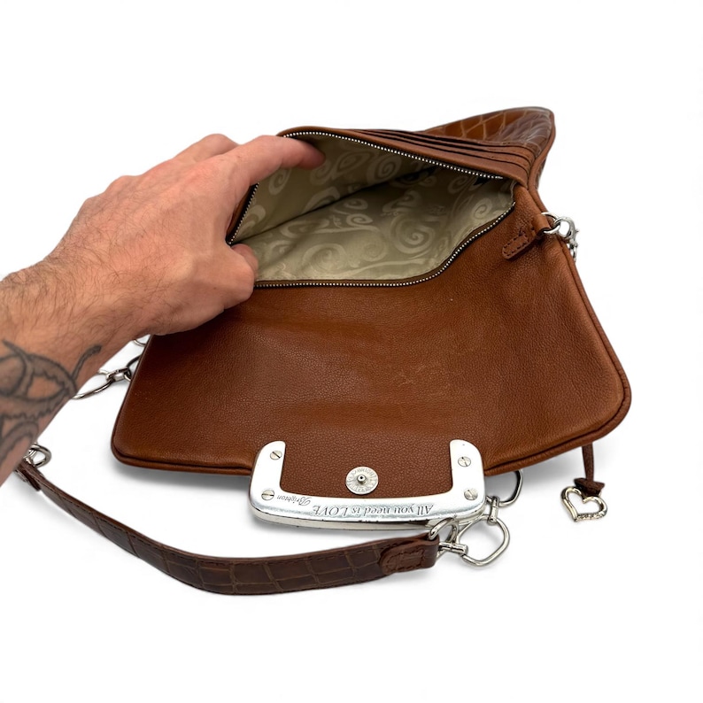 Puede incluir: Un bolso de cuero marr&oacute;n con un cierre plateado y una correa de cadena. El bolso est&aacute; abierto, revelando un forro interior estampado. El cierre dice "All you need is LOVE". Un peque&ntilde;o dije de coraz&oacute;n cuelga del lateral.