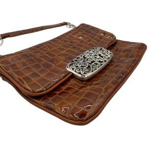 Puede incluir: Un bolso de mano marr&oacute;n texturizado con un cierre plateado. El bolso de mano tiene una correa larga y un cierre plateado rectangular con un dise&ntilde;o en espiral. El bolso est&aacute; hecho de un material brillante y tiene forma rectangular.