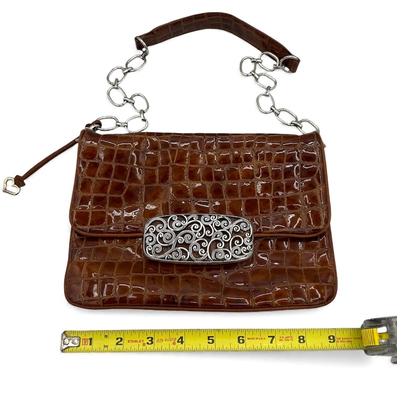 Puede incluir: Un bolso marr&oacute;n de piel de cocodrilo sint&eacute;tica con una correa de cadena plateada y un cierre decorativo plateado. El bolso tiene un cierre de solapa y un peque&ntilde;o dije en forma de coraz&oacute;n. El bolso mide aproximadamente 23 cm de ancho.