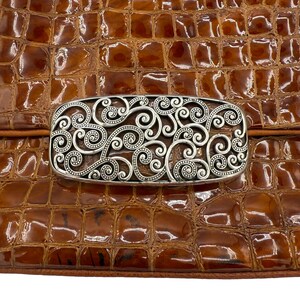 Puede incluir: Un bolso marr&oacute;n texturizado con un cierre rectangular plateado. El cierre presenta un dise&ntilde;o intrincado y arremolinado. La superficie del bolso tiene un patr&oacute;n similar al de un reptil, y el cierre est&aacute; centrado en la solapa del bolso.