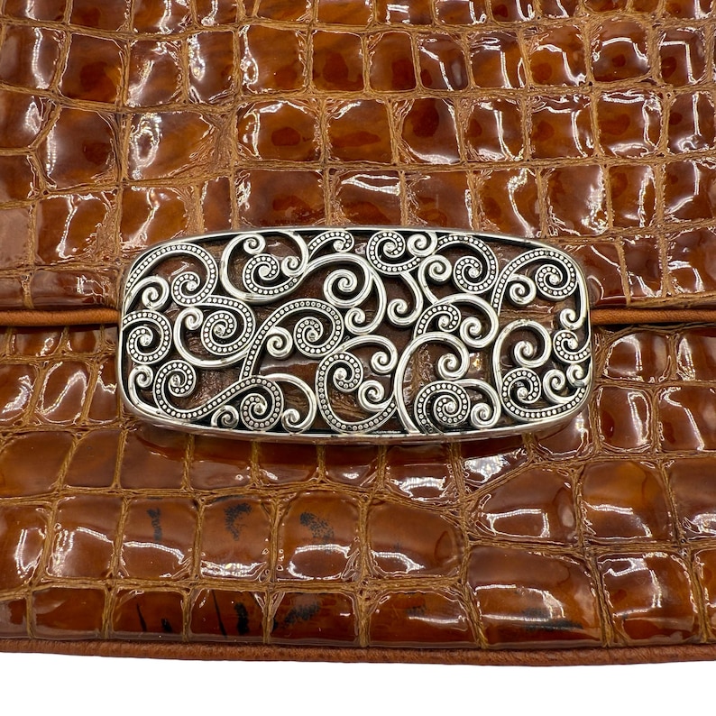 Puede incluir: Un bolso marr&oacute;n texturizado con un cierre rectangular plateado. El cierre presenta un dise&ntilde;o intrincado y arremolinado. La superficie del bolso tiene un patr&oacute;n similar al de un reptil, y el cierre est&aacute; centrado en la solapa del bolso.