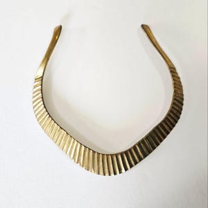 Könnte beinhalten: Ein goldfarbenes Halsband mit geripptem, strukturiertem Design. Die Halskette hat eine U-Form mit einer breiteren, strukturierten Mitte und glatten, gebogenen Enden. Das Metall hat eine warme, antike Oberfläche, und die Halskette ist vor einem weißen Hintergrund platziert.