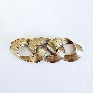 May include: A gold-toned metal brooch featuring a series of interlocking oval links. The brooch has a textured surface and a vintage aesthetic, suitable for adding a touch of elegance to any outfit.
