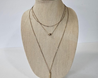 Puede incluir: Un conjunto de collares en capas exhibido en un maniquí beige. Los collares son de color dorado, con diferentes estilos y longitudes de cadena. Un collar presenta un colgante de barra vertical. El maniquí está sobre un fondo blanco.