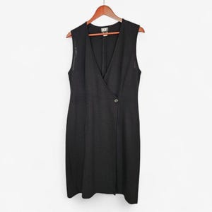 May include: Sleeveless black dress with a V-neckline and a wrap-style front. The dress features a single button closure and is hanging on a wooden hanger. The DKNY brand label is visible at the neckline.