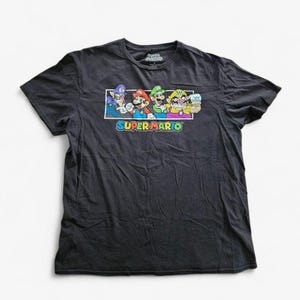 以下が含まれることがあります： スーパーマリオのキャラクター、ワリオ、マリオ、ルイージ、ワルイージのカラフルなグラフィックが特徴の濃いグレーのTシャツ。「スーパーマリオ」の文字がキャラクターの下にカラフルなフォントで印刷されています。柔らかい素材のシャツです。