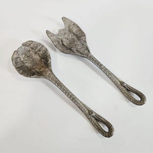 May include: A pair of silver-toned salad servers. One server is a spoon with an elephant head design, and the other is a fork. Both servers have textured handles and loop-shaped ends.