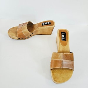 May include: A pair of tan leather slide sandals with wooden wedge heels and soles. The wide leather straps feature visible stitching and are secured to the wooden base with small metal studs. The brand name l.e.i. is printed on the heel bed.