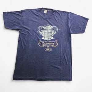 May include: Navy blue t-shirt featuring a vintage car graphic. The graphic includes the word "Duesenberg" in a decorative font, with "Model J" below. The shirt has short sleeves and a crew neck.