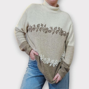 May include: A beige turtleneck sweater with a floral design. The sweater has a two-tone design with a lighter beige top and sleeves, and a darker beige bottom. The floral design is embroidered in brown and white.