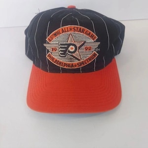 Op de afbeelding: Een vintage honkbalpet in zwart en oranje, met een gebogen klep. De pet heeft witte krijtstrepen en een geborduurd embleem met de tekst "43rd NHL All-Star Game 1992 Philadelphia Spectrum". Het heeft een oranje klep en een kleine oranje knop aan de bovenkant.