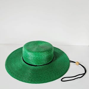 Può includere: Un cappello verde brillante con una tesa larga e una corona cilindrica. Il cappello ha una fascia nera attorno alla corona e un sottogola nero con una perla di legno. Il cappello è realizzato in materiale tessuto.