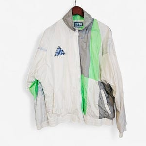 Op de afbeelding: Een vintage windjack in wit, grijs en groen met een volledige ritssluiting. De jas heeft een colorblock-ontwerp, een driehoekig logo op de borst en de tekst "H.I.C." op de kraag.