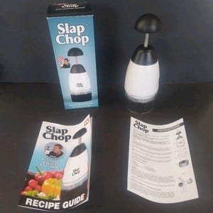 May include: A white and black Slap Chop food chopper with a black handle and clear base. The product is displayed with its box, recipe guide, and instructions. The box and recipe guide feature the product name and images.