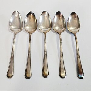 May include: Five silver spoons arranged in a row. Each spoon has an oval bowl and a long, slender handle with a decorative floral pattern near the top. The handles have a geometric, faceted design at the base.
