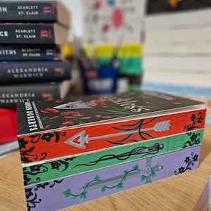 Può includere: Una pila di libri con bordi dipinti. Il libro in alto ha una copertina nera con disegni floreali rossi e il titolo "Ruthless". I bordi sono dipinti di rosso, verde e viola, ognuno con un design diverso: una freccia, una spada e delle viti.