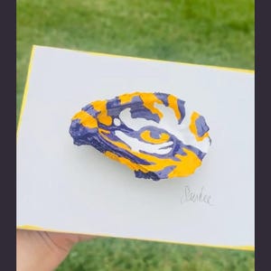 May include: A white rectangular card featuring a hand-painted oyster shell with a tiger eye design in vibrant purple, gold, and white paint. The shell is centered on the card, which displays the handwritten signature Sandee in the bottom right corner.