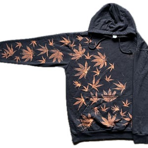 Op de afbeelding: Een donkergrijze hoodie met een voorzak en een capuchon. De hoodie heeft een patroon van oranje cannabisbladeren over de voorkant en de linkermouw. De bladeren zijn in verschillende maten en oriëntaties.