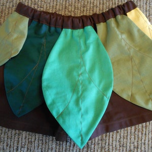 May include: A brown skirt with green and yellow leaf-shaped fabric panels sewn onto it. The skirt is made of a soft, brown fabric and has a simple design.