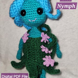May include: A handmade crochet doll, Nadia the Nymph, with blue hair and teal skin. The doll wears a green dress adorned with pink flowers. The text "Nadia The Nymph" and "Digital PDF File" are visible.
