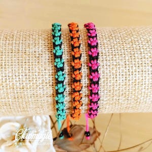 Puede incluir: Tres pulseras de macramé hechas a mano en verde azulado, naranja y rosa, cada una con un acento negro. Las pulseras se exhiben sobre una superficie texturizada. Las pulseras tienen un diseño de huella de pata.
