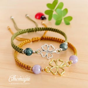Könnte beinhalten: Zwei Makramee-Armbänder mit Kleeblatt-Anhängern und Edelsteinperlen. Eines hat einen silbernen Kleeblatt-Anhänger und grüne Perlen, das andere einen goldenen Kleeblatt-Anhänger und lila Perlen. Auf einer Holzoberfläche.