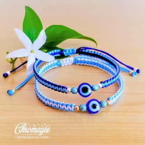Peut inclure: Deux bracelets macramé bleus et blancs avec un charme d'œil de protection. Les bracelets sont présentés sur une surface en bois, avec une fleur blanche et des feuilles vertes en arrière-plan. Les bracelets ont des cordons réglables.