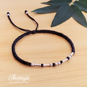 Puede incluir: Pulsera trenzada negra con cuentas y detalles plateados. La pulsera tiene cordones negros ajustables y se muestra sobre una superficie de madera clara. La palabra "Chromagie" es visible en la esquina inferior izquierda.