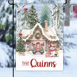 May include: A winter-themed garden flag featuring a snow-covered cottage with a wreath on the door and the text "The Quinns" in red. A snowman and deer are in front of the house. The flag is decorated with Christmas trees and red bows.