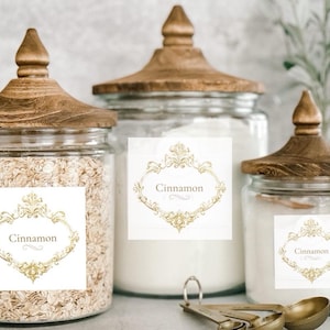 May include: Three glass jars with wooden lids, labeled "Cinnamon" in an ornate gold frame. One jar contains oats, another white powder, and the third a similar white substance. A set of measuring spoons is in the foreground.