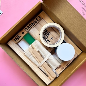 May include: A craft kit in a cardboard box containing a wooden screen printing frame with the text INK + ORANGES, a roll of masking tape, a small jar with a white lid, two wooden stir sticks, a piece of fabric, and a green rectangular block.