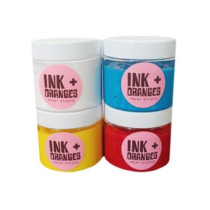 May include: Four jars of paint, each with a white lid and a pink label that says "INK + ORANGES PRINT STUDIO". The jars are filled with white, blue, yellow, and red paint.